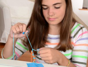 A pretty girl macrames a colorful friendship bracelet using blue and orange thread.